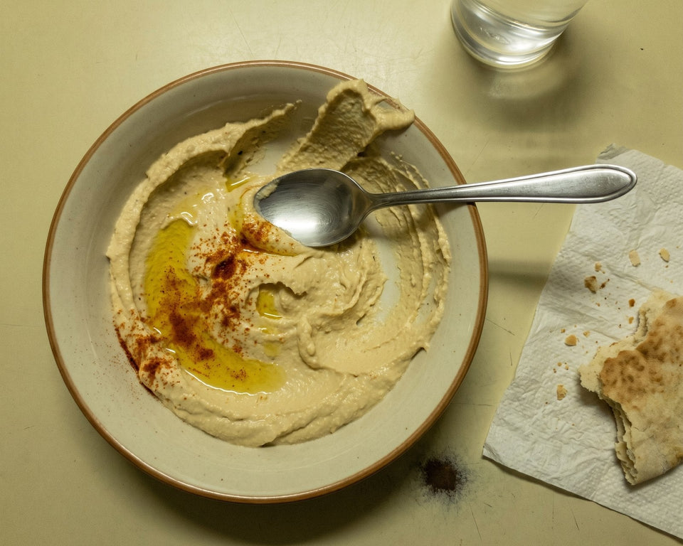 Uma tigela de hummus com azeite e páprica, uma colher e pão sírio.