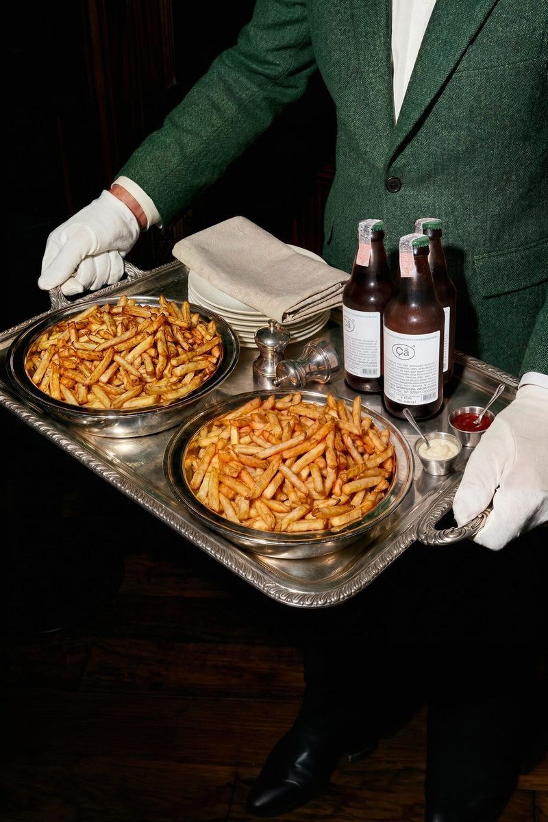 Garçom de terno verde leva uma bandeja com batatas fritas e garrafas de Çã