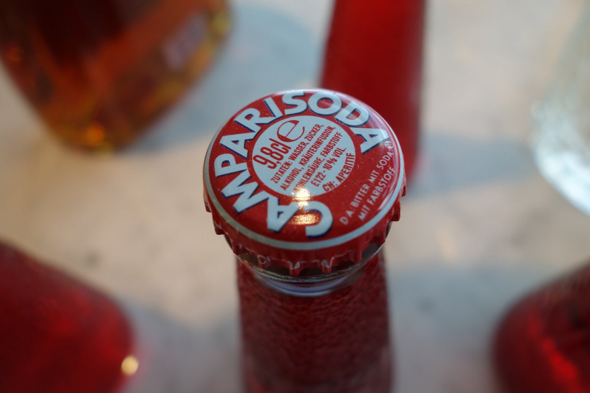 Close da tampinha vermelha do Campari Soda, com o nome gravado em volta, e outras garrafinhas fora de foco atrás.