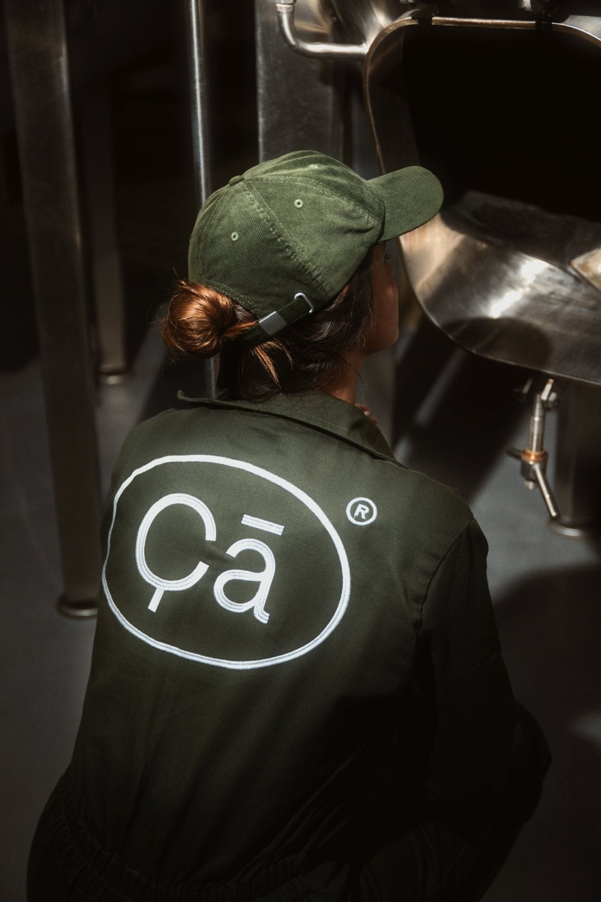De costas, uma pessoa de boné e macacão verdes, com o logo da Çã bordado nas costas, diante de um tanque de inox
