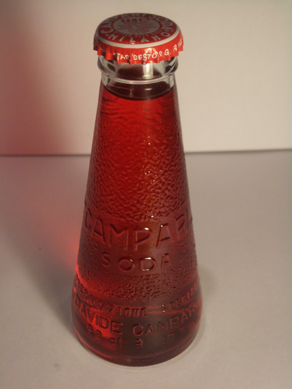 Uma garrafinha de Campari Soda sobre fundo claro; o nome aparece em relevo no próprio vidro.